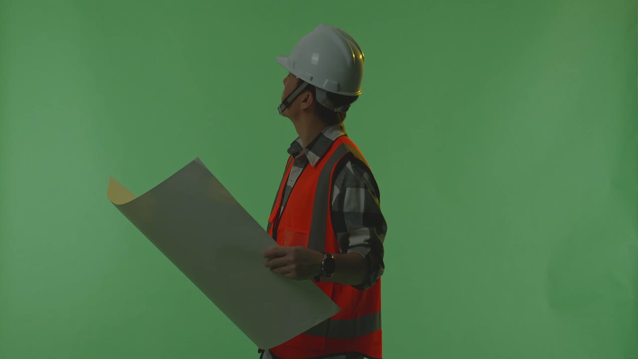 vista lateral de un ingeniero masculino asiático con casco de seguridad mirando el plano en sus manos y mirando a su alrededor mientras está de pie en el estudio de fondo de la pantalla verde, durante la puesta o el amanecer