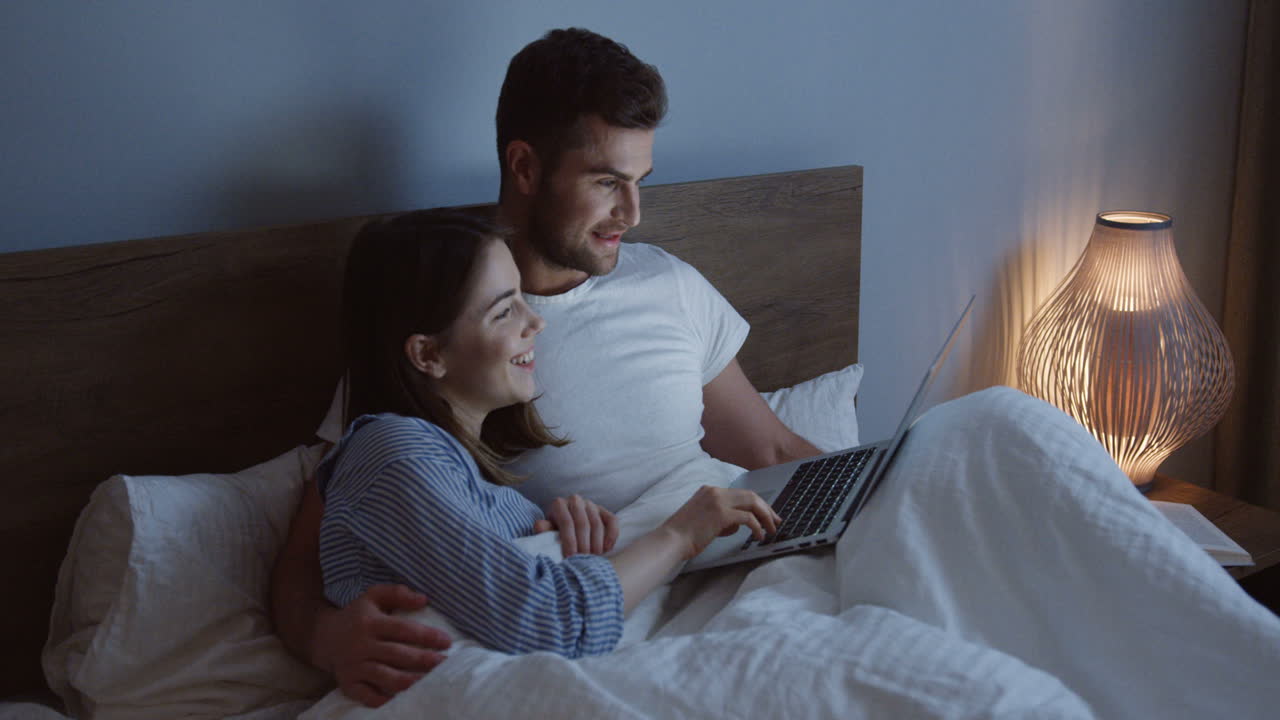 sonriente pareja joven caucásica de buen aspecto viendo algo divertido en la computadora portátil en la cama por la noche