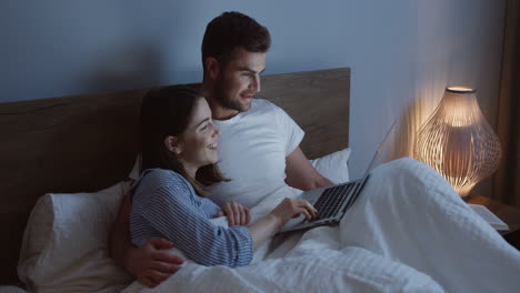 smiling good looking caucasian young couple watching something funny on the laptop in the bed at night