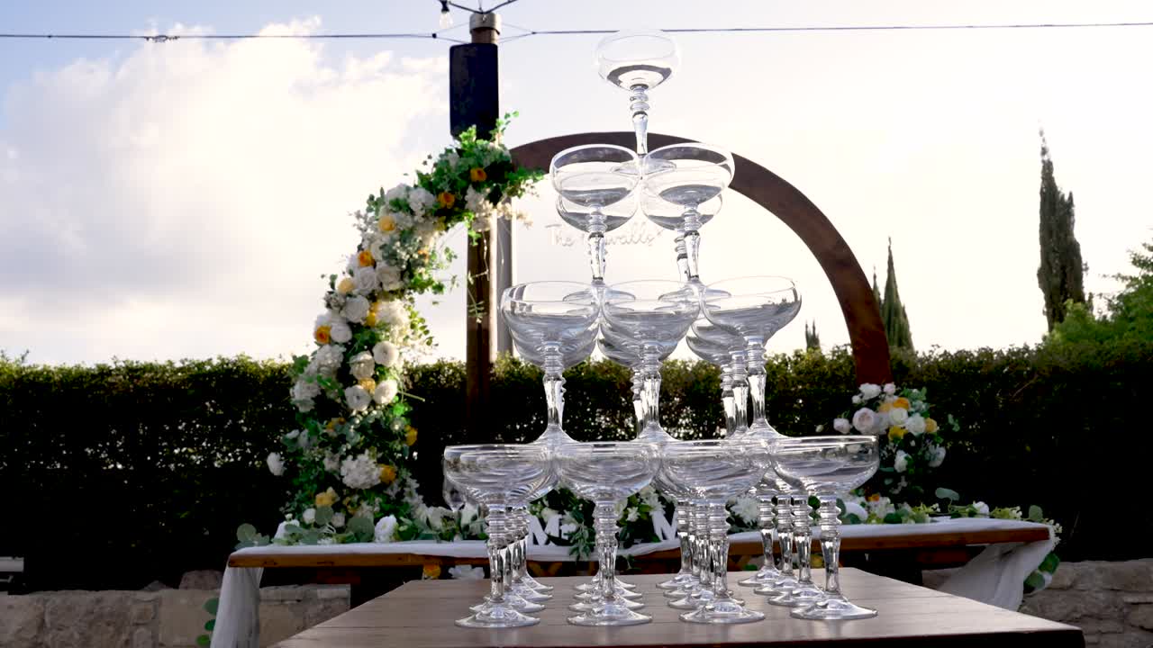 Una splendida composizione di bicchieri impilati attende gli ospiti in una pittoresca ambientazione esterna, ornata di fiori e vegetazione. Questo elegante allestimento crea l'atmosfera perfetta per un'occasione speciale.