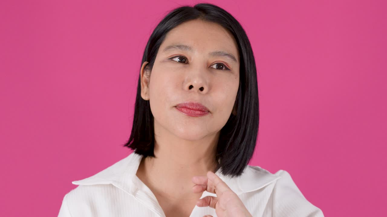 Cảnh quay trung bình một người phụ nữ châu Á trầm tư chạm vào tóc cô ấy trên nền màu hồng rực rỡ.