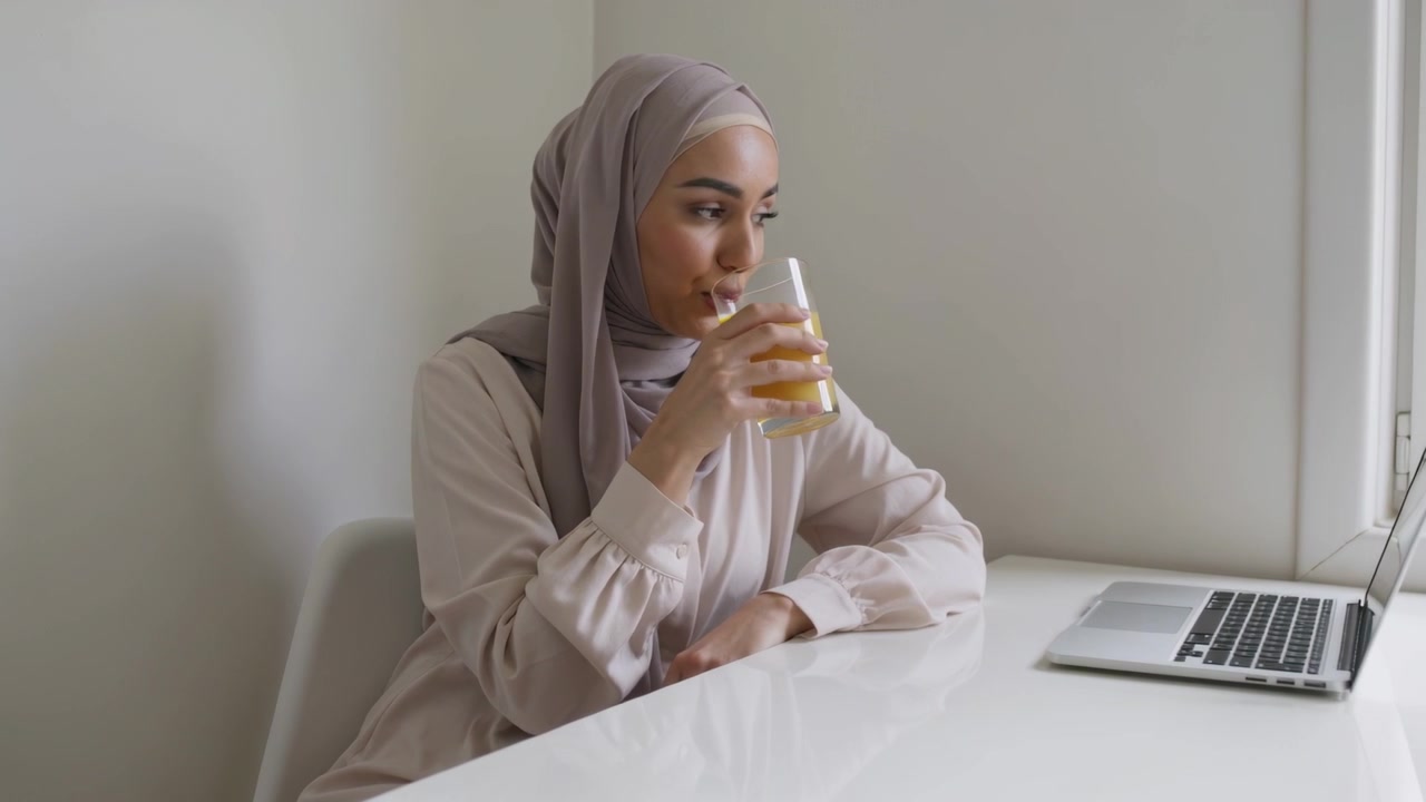 Smiling woman wearing hijab lifting glass to lips at home desk, checking laptop while working