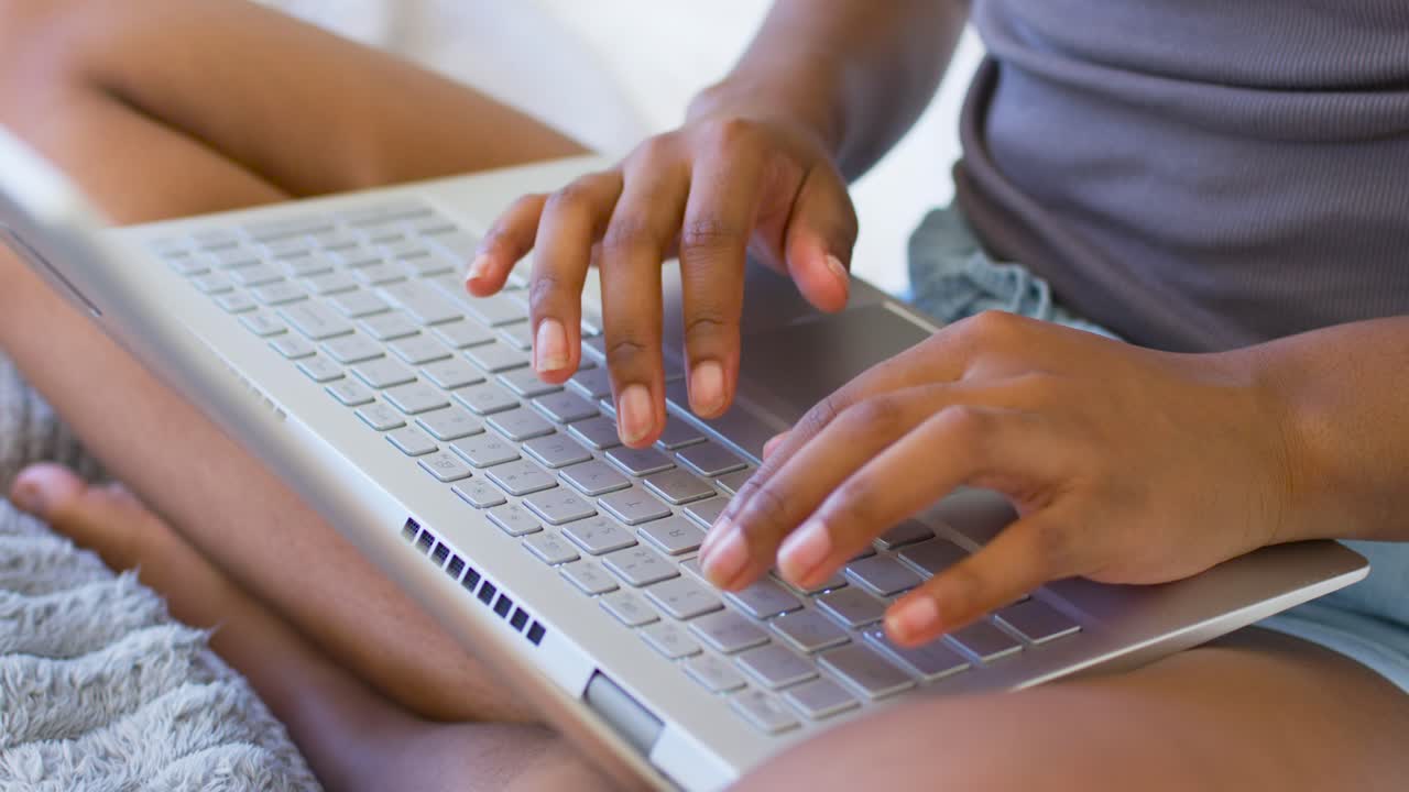 Female placing hands on keyboard, typing on silver laptop on bed in tank and shorts working