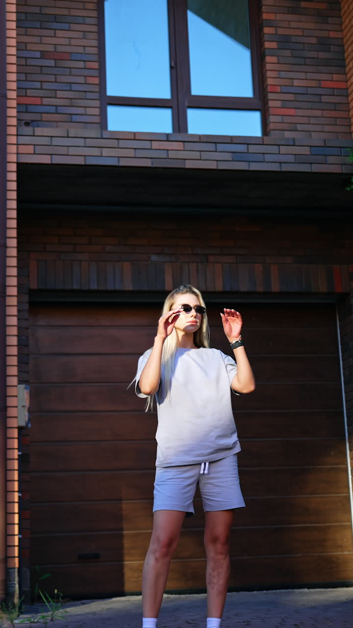 Blonde lady wearing sunglasses stands outdoors holding hands in the pockets. Low angle view at the model demonstrating sportswear. Vertical video