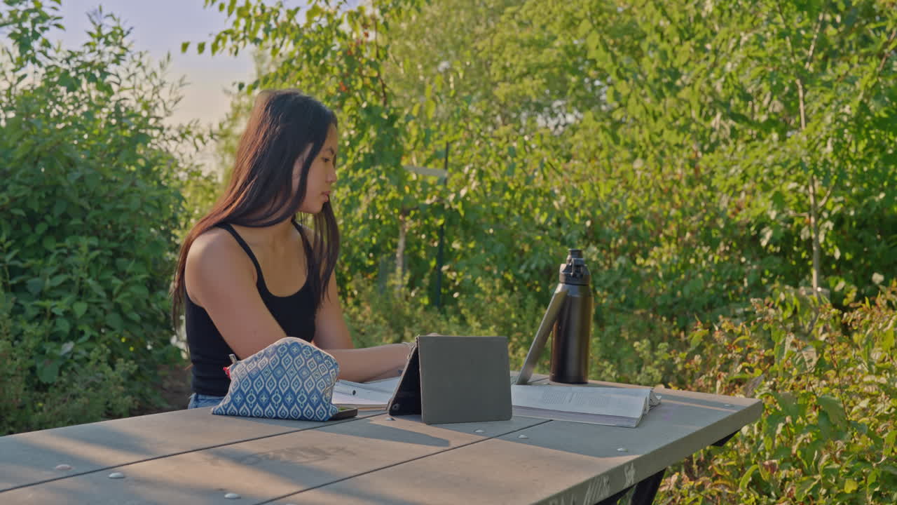 Young Asian student sitting at a park table with tablet, notebook and water bottle, studying outdoors, dolly shot moving right, focus on education and sustainability