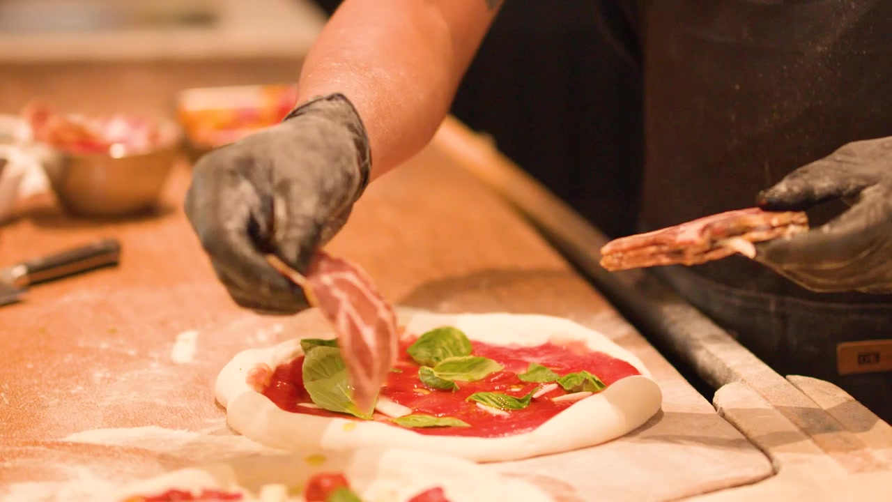 Chef professionista aggiunge fette di salume all'impasto della pizza con salsa di pomodoro e foglie di basilico.