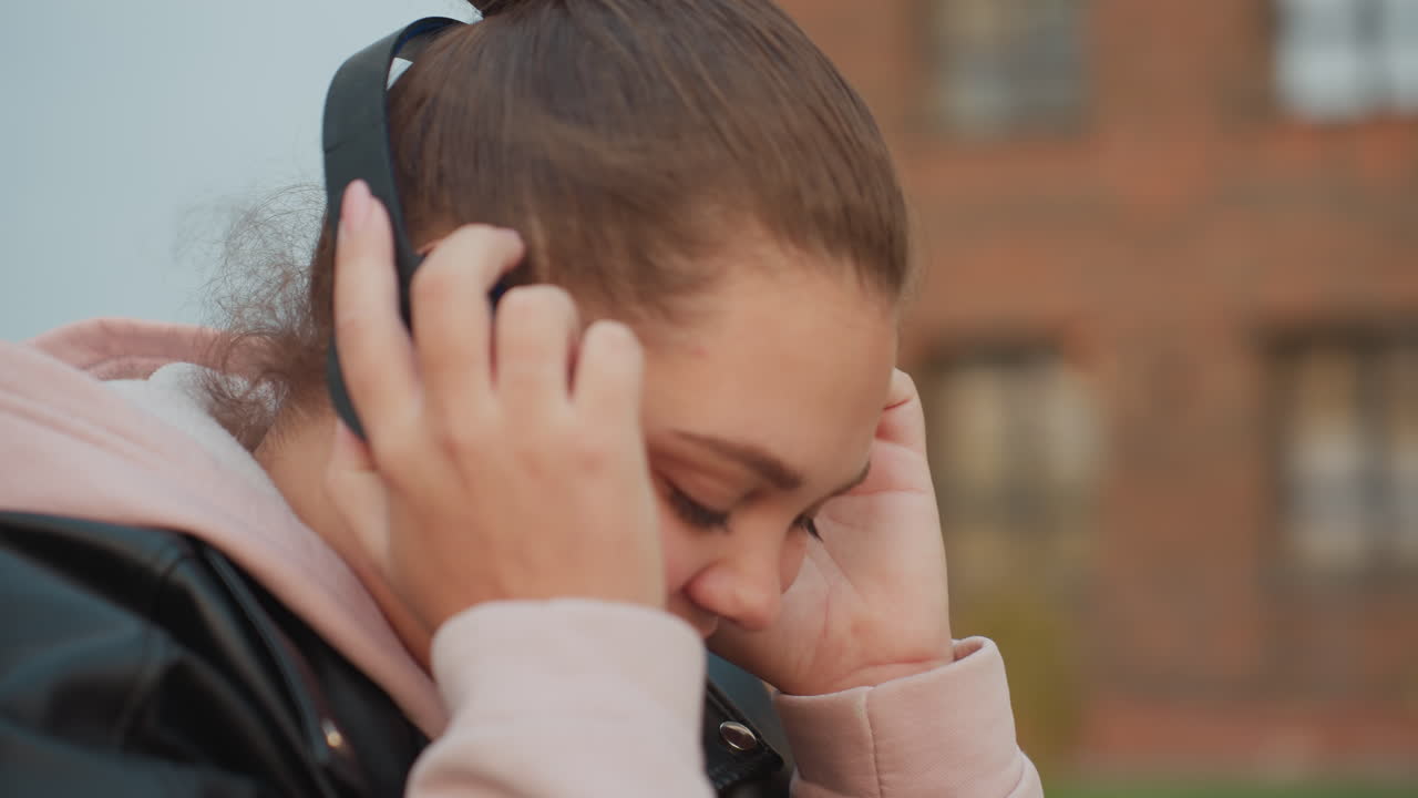 Close up young woman in pink hoodie and leather jacket gently puts on headphone while enjoying cool morning breeze with soft background bokeh and blurred building in view