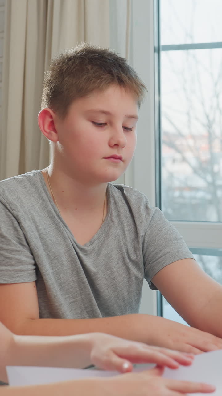 Ragazzo che piega la carta con concentrazione, preadolescente caucasico che pratica origami precisi a un tavolo di vetro, maglietta grigia, espressione calma, mani ferme, la ripetizione e la pazienza sviluppano l'abilità, interno silenzioso