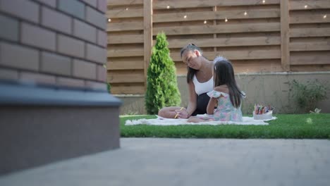 a mother and her young daughter sit on a blanket in the backyard, engaging in a drawing activity together. the outdoor scene captures a peaceful family moment, fostering creativity and bonding.