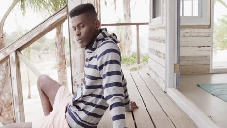 african american man sitting on steps of wooden beach house relaxing in the sun, slow motion