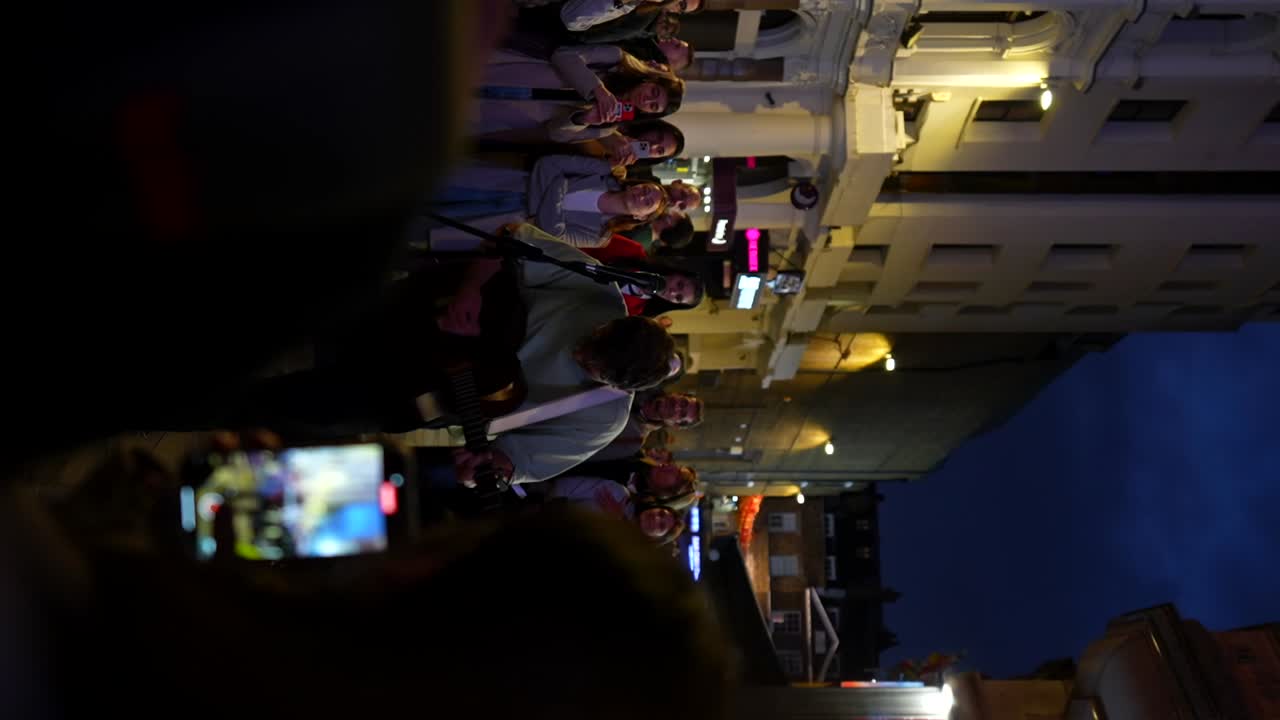 Crowd gathers at night around a street musician in Leicester Square for a lively group singalong, London. Vertical