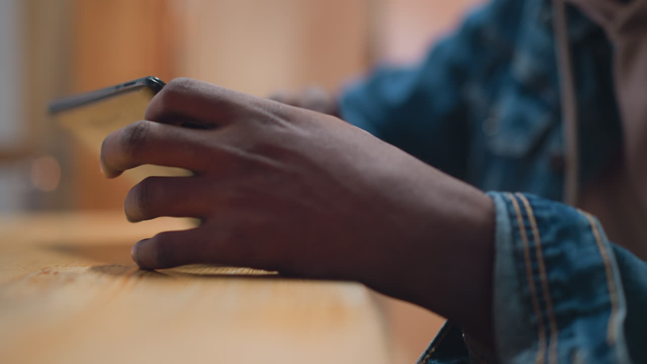 close up black hand in denim sleeve holding tablet on wooden table in cozy cafe with soft bokeh and warm ambient lighting conveying modern digital leisure moment