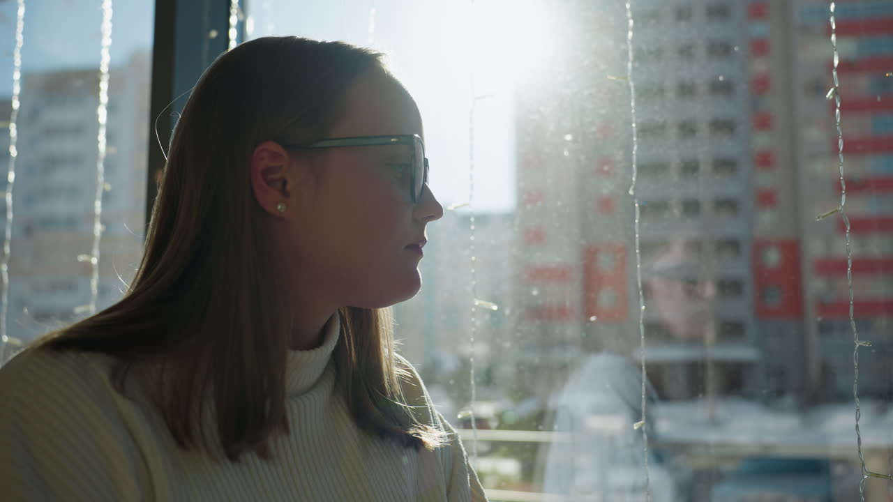 Calm artist sits beside large glass panel, gazing upward with thoughtful expression as sunlight floods cafe interior, city buildings blur behind frosted glass