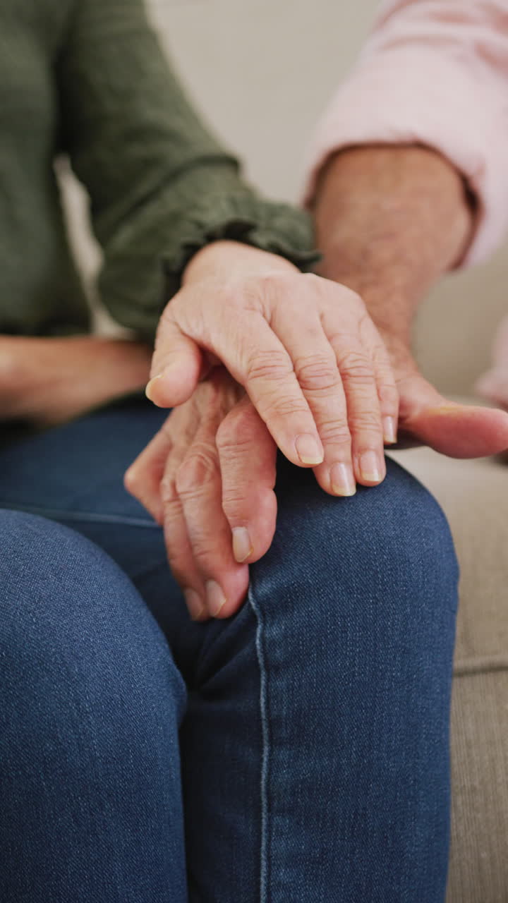 Vertical video of diverse senior couple sitting on couch and holding hands