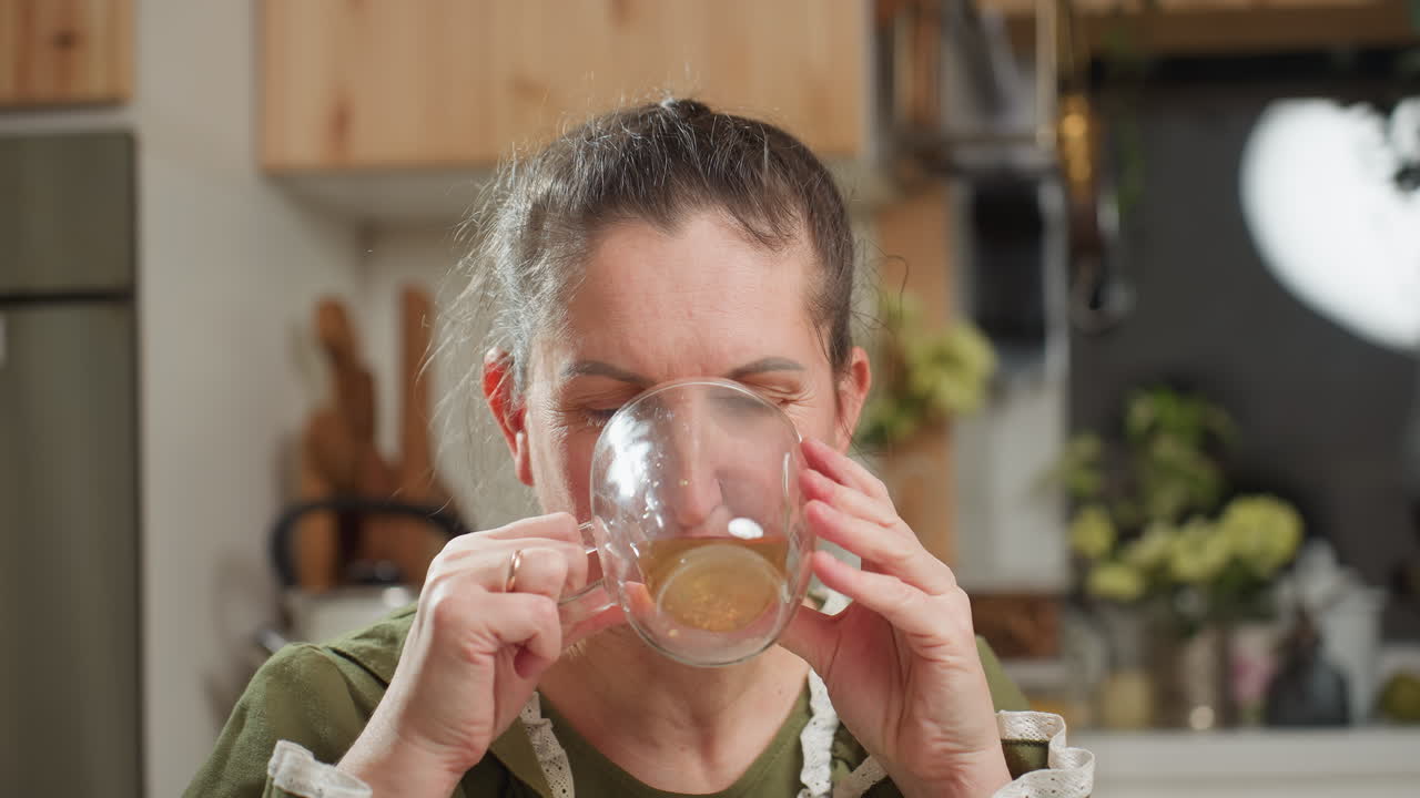 White lady drinks coffee from transparent glass cup in cozy kitchen setting, then lowers cup slowly while gazing calmly at camera with relaxed expression