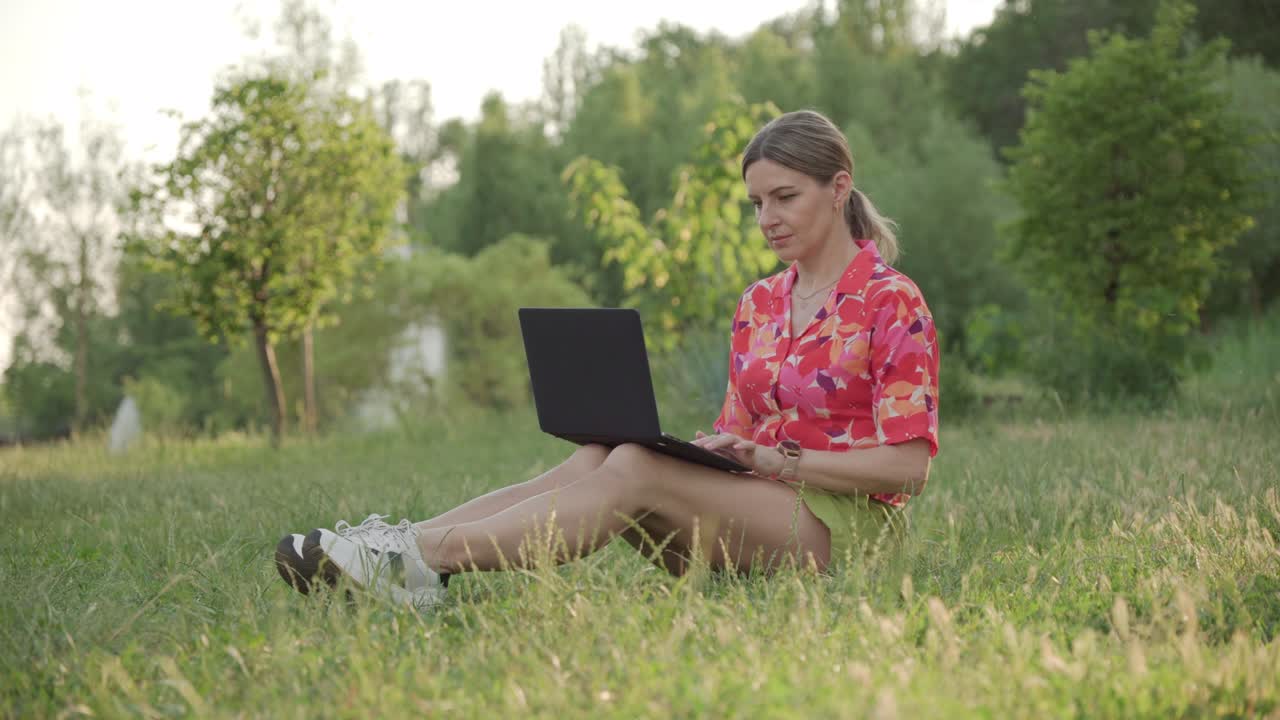 Business woman working remotely in the park using a laptop. She stretches and straightens her back from fatigue.