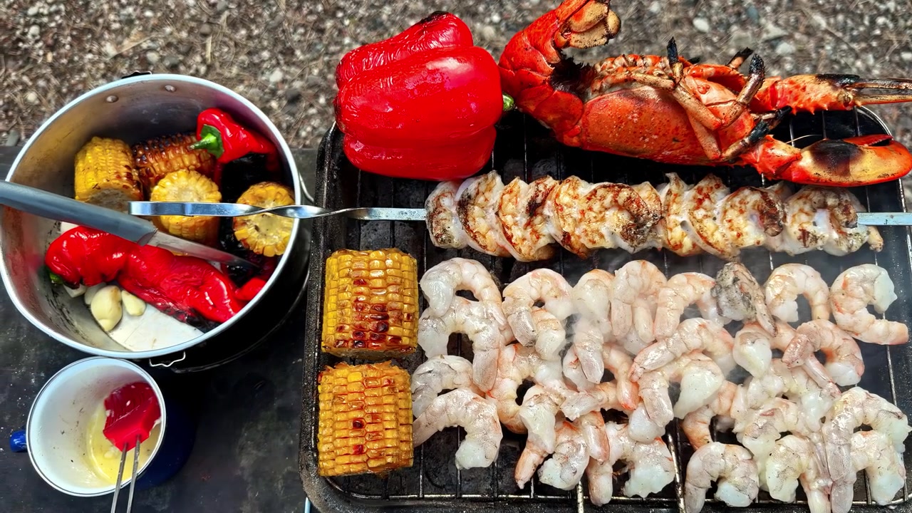 Ovenfra-billede af hel grillet hummer, rejespyd, majs og rød peber, der tilberedes på en udendørs grillrist ved siden af en gryde med ristede grøntsager.