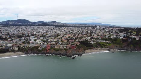 Imágenes-Aéreas-De-Drones-De-4.000-Fps-De-La-Costa-De-California-Cerca-De-La-Ciudad-Y-La-Playa-De-Presidio:-Olas-Rompiendo-Contra-Los-Acantilados-De-La-Costa,-Playa-De-Arena,-Cielo-Nublado,-Olas-Blancas-Y-árboles-De-Hoja-Perenne-Salpican-El-Paisaje