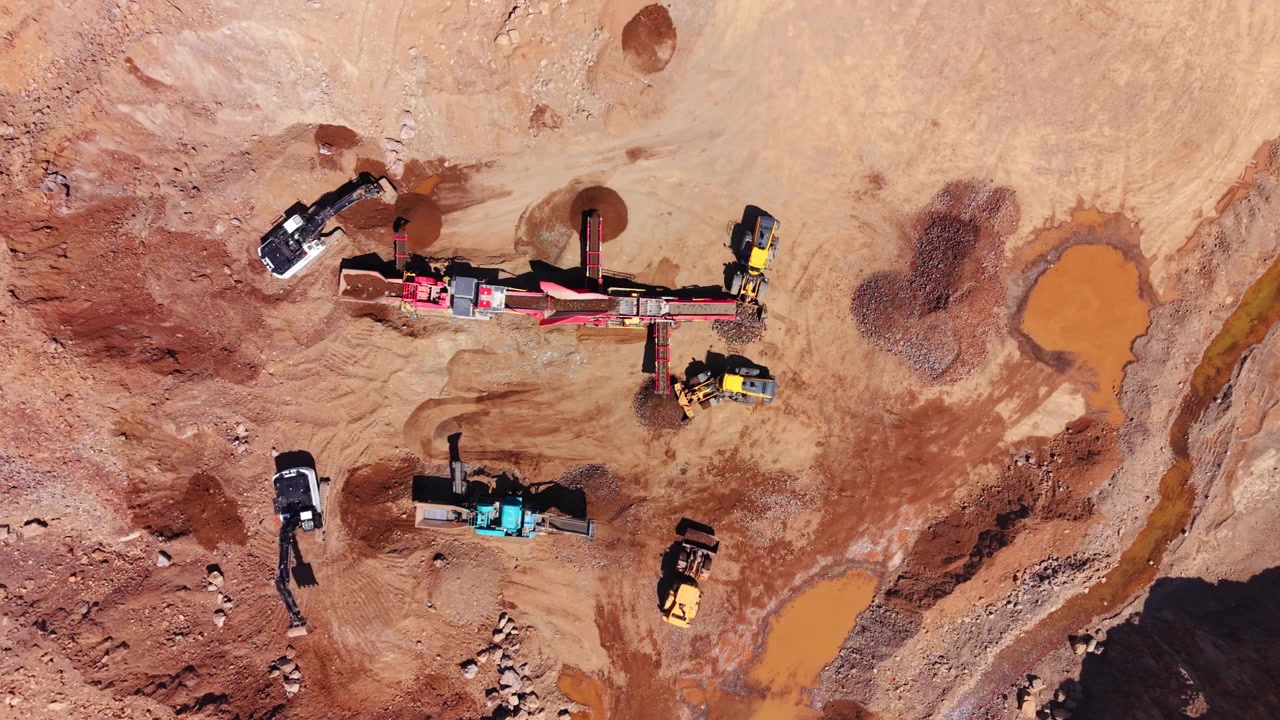 Aerial view of crushing plant and dump trucks at mine site. Red machinery, excavators and orange soil ponds below.