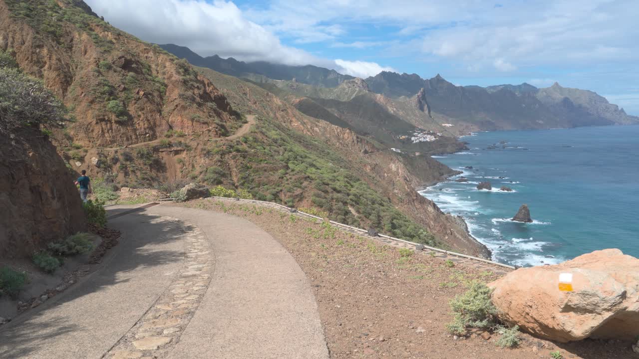 pareja caminando por una carretera costera con impresionantes vistas, alta línea montañosa, cielo azul claro, espesas nubes blancas y olas rompiendo