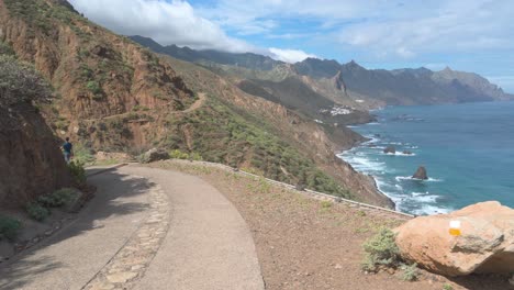 Pareja-Caminando-Por-Una-Carretera-Costera-Con-Impresionantes-Vistas,-Alta-Línea-Montañosa,-Cielo-Azul-Claro,-Espesas-Nubes-Blancas-Y-Olas-Rompiendo