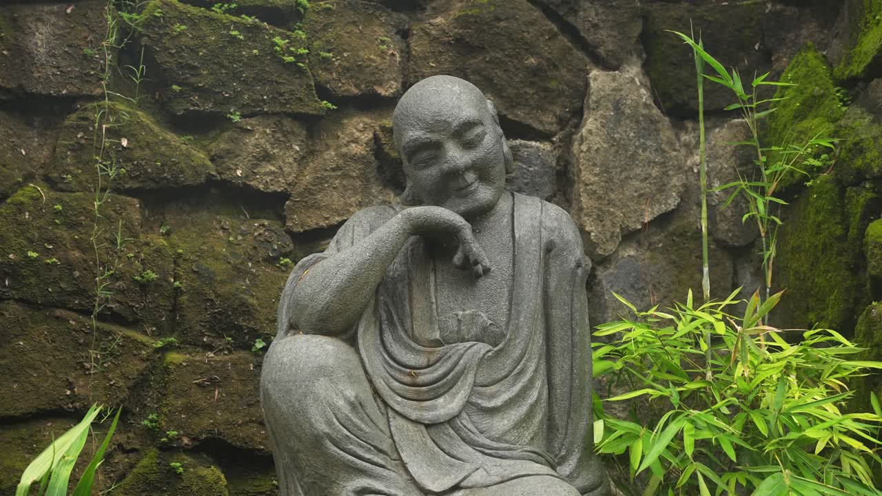 A close-up centers on the contemplative stone figure, head tilted and hand to cheek. Mossy wall and bamboo fade behind, spotlighting texture, serenity, and quiet presence in the natural frame