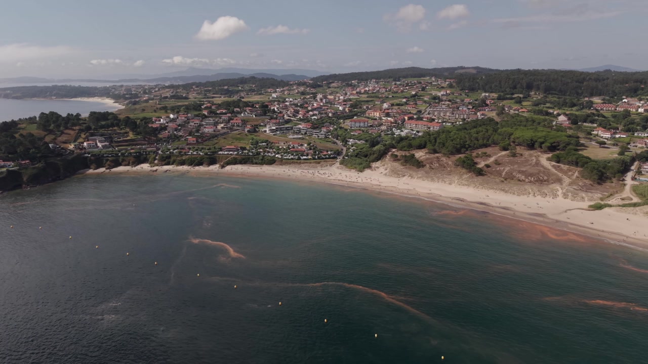 Roterende luftfoto fra drone over Montalvo-bugten, der viser en buet sandstrand, turkist vand, grønne klitter og en kystby i Galicien, Spanien