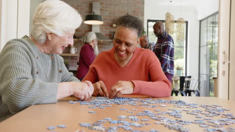 Happy-diverse-senior-female-friends-talking-and-doing-jigsaw-puzzle-at-dining-table,-slow-motion