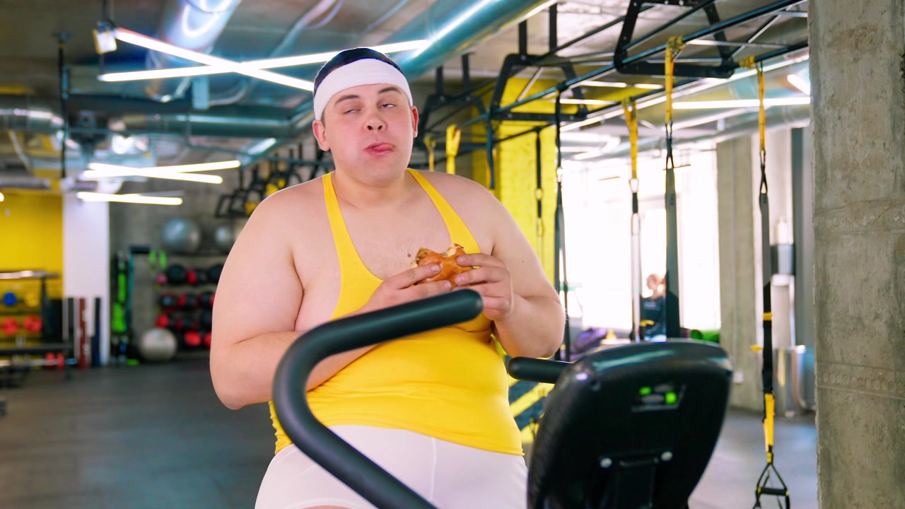 Overweight Man Eating a Burger on an Exercise Bike in a Gym