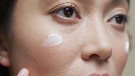 asian woman with dark hair applying cream on her face on beige background, slow motion
