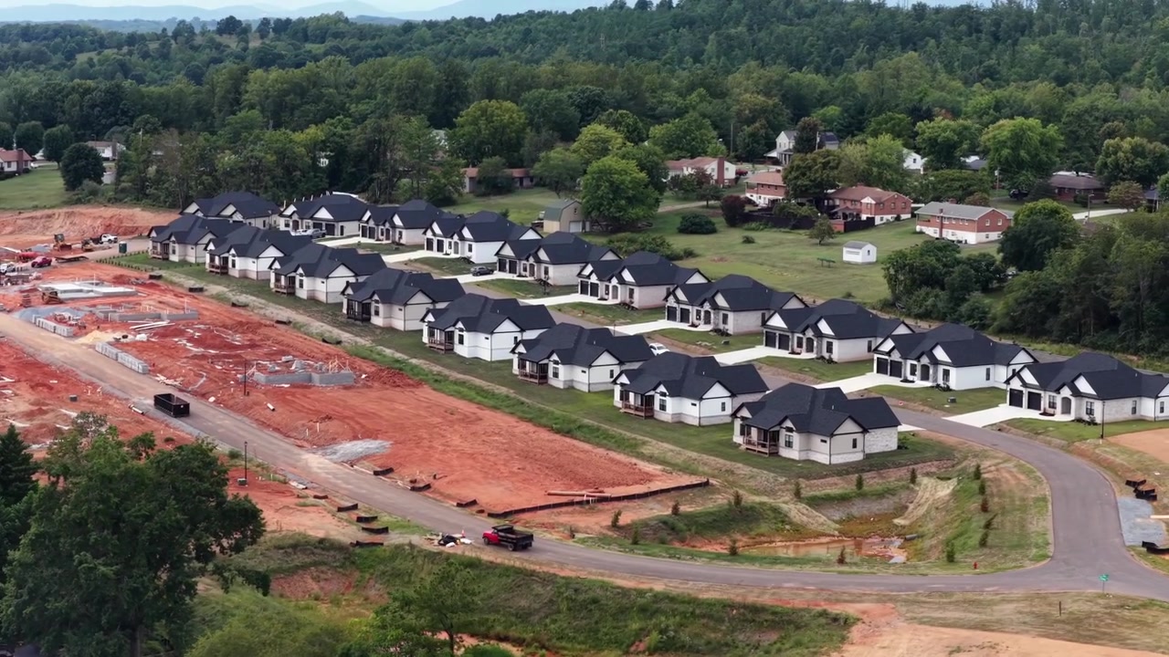 New white houses with black roofs stand in tight rows above raw red clay lots on a growing residential construction site outside Lynchburg, Virginia. Aerial lateral wide shot