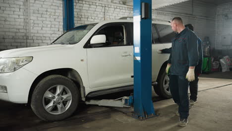 mecánicos en uniformes azules inspeccionando y observando un automóvil que se levanta en un taller, centrándose en los detalles del vehículo, con equipos y herramientas visibles en el fondo