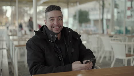 man using smartphone and smiling at camera in outdoor cafe