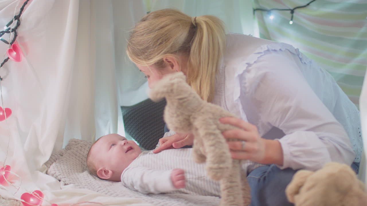 Loving mother playing game with  baby son and soft toy lying on rug in homemade camp in child's bedroom at home - shot in slow motion
