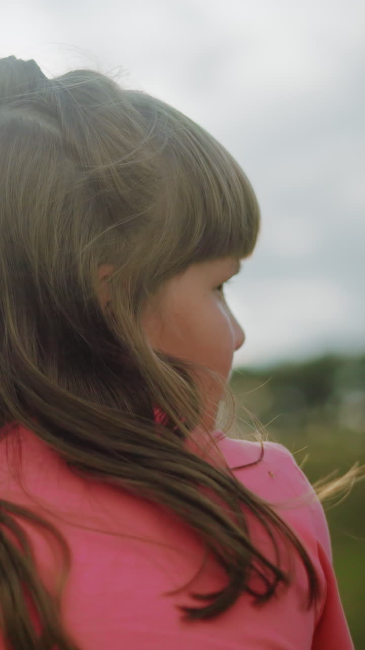 vista trasera de una mujer de color rosa con el cabello largo y ondulado que fluye en el viento, iluminado por la luz del sol, el suave resplandor natural crea una atmósfera de campo de ensueño con árboles borrosos y cielo abierto en el fondo
