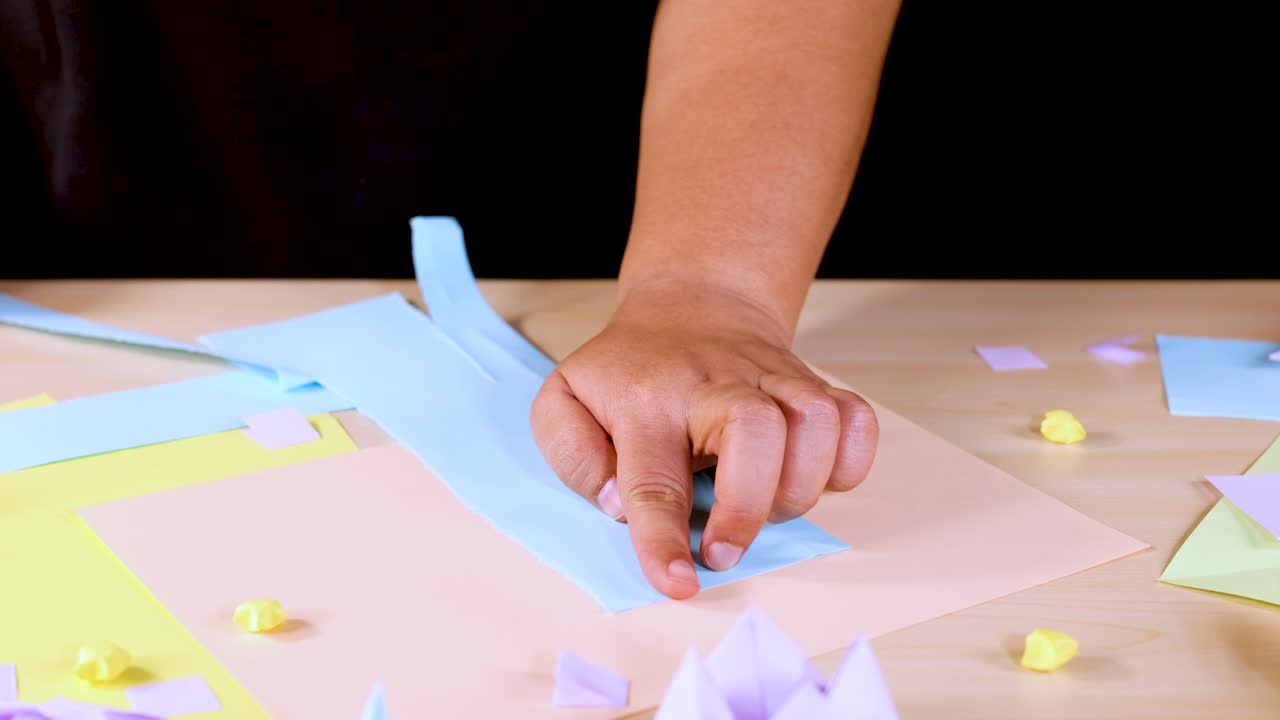 Person tears blue paper strip on desk with pastel sheets, origami, and soft lighting