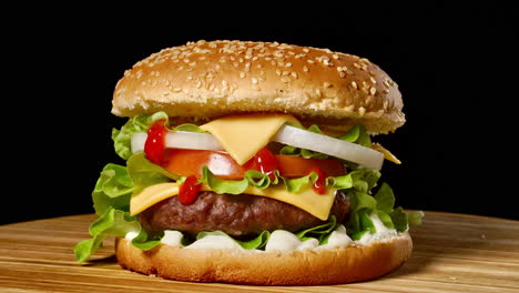 gran hamburguesa con chuleta de carne, tomates, hongos y pepinos con queso derretido gira en una tabla de madera sobre fondo negro.