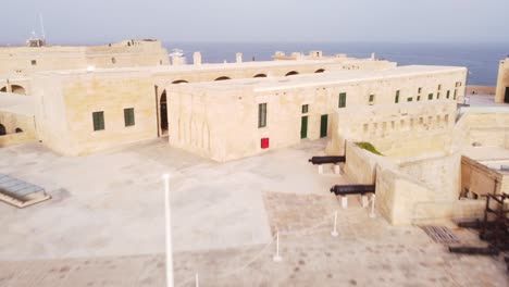 Close-up-drone-flight-over-heavy-armed-medieval-fortress-of-Valletta-Malta-out-to-view-the-scenic-open-sea-with-a-bridge-and-boats