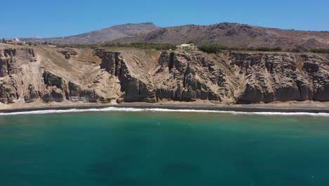 aerial flying backwards of greek island, rocky cliff and blue water waves in the mediterranean sea on white beach in santorini, greece