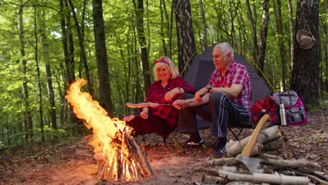 Senior-ältere-Großmutter-Großvater-Kochen-Braten-Würstchen-über-Lagerfeuer-Im-Holz-Beim-Camping