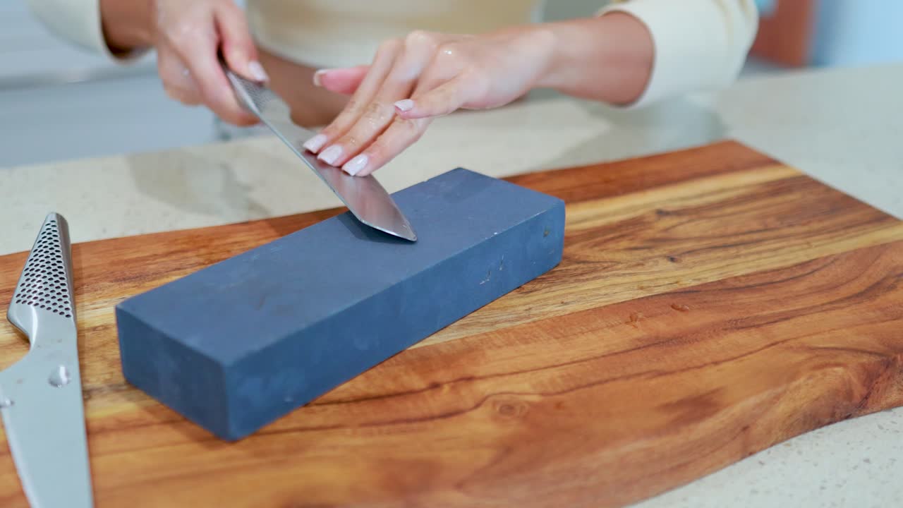 A person sharpens a stainless steel chef’s knife using a large whetstone on a wooden cutting board under bright, natural kitchen lighting with steady camera framing