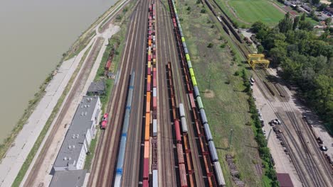 aerial view of a large rail yard with multiple freight trains carrying colorful cargo containers. the facility is located beside a river, surrounded by green fields and industrial buildings