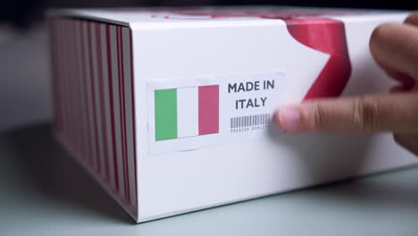 Manos-Aplicando-Etiqueta-De-Bandera-Hecha-En-Italia-En-Una-Caja-De-Cartón-De-Envío-Con-Productos