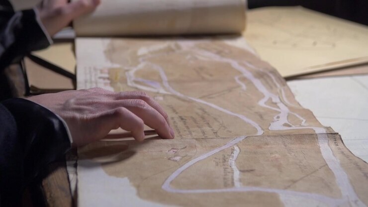 Close-up of a hand examining an antique map spread across a wooden table. Historical cartography, exploration, navigation, geography and archival research concept.