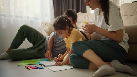 Der-Glückliche-Junge-Familienvater-Zeichnet-Zusammen-Mit-Seiner-Tochter-Und-Seiner-Frau-Mit-Bunten-Bleistiften-Auf-Papier,-Während-Er-In-Einem-Modernen-Zimmer-Auf-Dem-Boden-Neben-Dem-Sofa-Sitzt