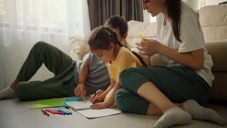 un joven de familia feliz junto con su hija y su esposa dibujan con lápices de varios colores en papel mientras están sentados en el suelo cerca del sofá en una habitación moderna