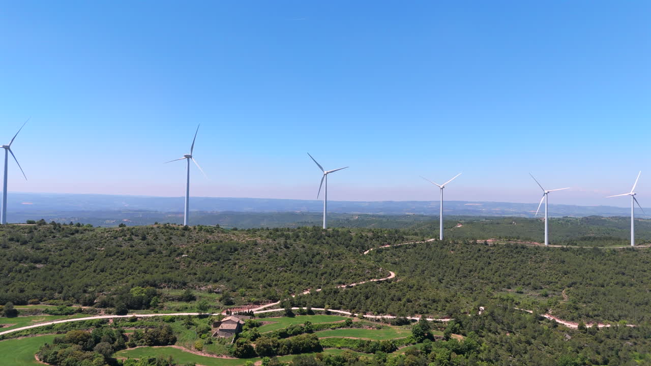 Drone flies forward toward a spinning wind turbine on a lush hilltop. Curved dirt road winds through green landscape under clear blue sky, with distant hills completing the panoramic view