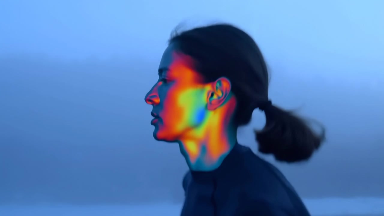 Side view follows a woman jogging steadily through a cold foggy setting, her ponytail moving with each stride while bright rainbow light patterns illuminate her face.