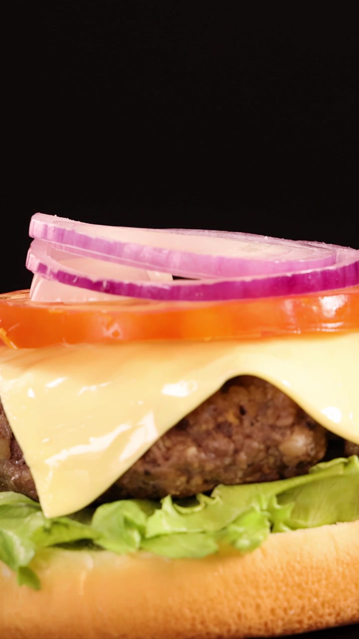 Hands carefully add tomato, onion, and a sesame seed bun to a cheeseburger with lettuce and cheese, under bright studio lighting and a black background