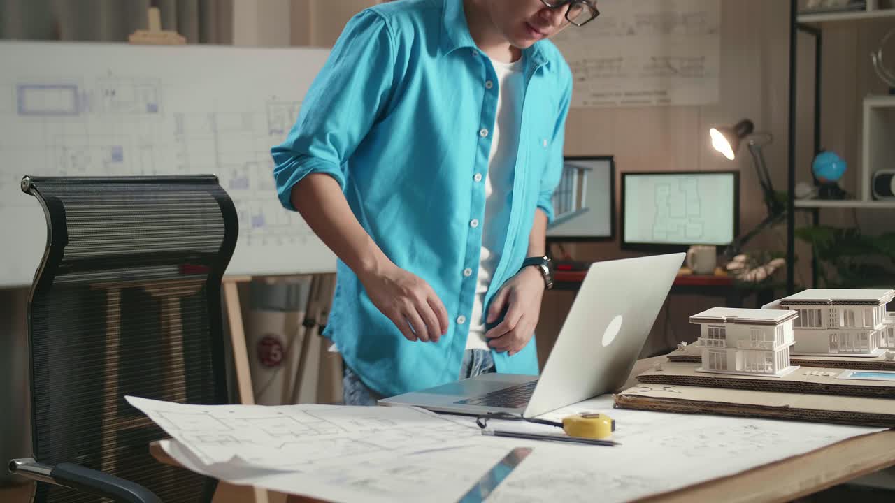Asian Man Engineer Walk Into The Office Before Typing On A Laptop And Looking At Blueprint